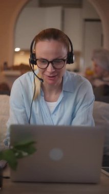 Mavi gömlekli, kulaklıklı, dizüstü bilgisayarlı ve akıllı telefonlu, rahat bir oturma odasında rahat bir şekilde oturan, yumuşak ışıklandırma, bitkiler ve arka planda görünen rahat bir atmosferi olan beyaz bir kadın.