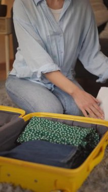 Açık mavi gömlekli kadın, arka planda bir kanepe ve dekoratif eşyalarla rahat bir oturma odasında yumuşak bir halının üzerinde diz çökerken sarı bir bavula giysilerini koymaya devam ediyor.