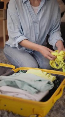 Açık mavi gömlekli kadın sarı bir bavulun içine dikkatlice kıyafet yerleştirir, katlanmış eşyaları düzeltir ve rahat bir yerde yumuşak bir halının üzerinde otururken yaklaşan yolculuğa hazırlanır.