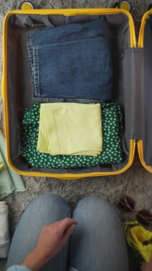 Kadın sarı ve yeşil giysileri sarı bir bavulun içine yerleştirir, katlanmış kot pantolonları ve gömlekleri, kapalı bir ortamda yumuşak, dokulu bir yüzeye dikkatlice yerleştirir.