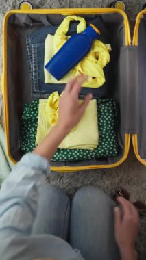 Kadın yolculuk için sarı bir bavul hazırlıyor, içine sarı gömlekler, kot pantolon ve hasır şapka yerleştiriyor, yanında mavi bir şişe ve kulaklık,