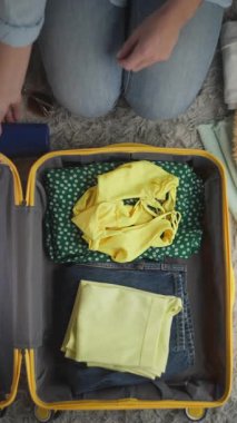 Kadın dikkatlice katlanmış giysiler, güneş gözlükleri ve aksesuarları sarı bir bavula yerleştiriyor, rahat bir kapalı alanda yumuşak ışıklandırma ve rahat bir halı ile organize bir paketleme işlemi gösteriyor.