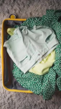 Renkli yaz kıyafetleri ve aksesuarları sarı bir bavula yerleştirilir. Yeşil benekli kumaş, hasır şapka ve mavi su şişesi sergilenir. Tatil ve seyahat hazırlıkları için idealdir.