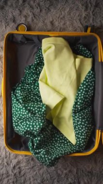 Yeşil benekli elbise, sarı şapka ve açık mavi gömlek gibi renkli eşyalar yumuşak, dokulu bir yüzey üzerinde açık sarı bir bavul içinde düzenlenir, canlı bir paketleme süreci vurgulanır