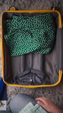 Kadın, yeşil benekli elbise ve sarı bluz da dahil olmak üzere çeşitli giysi parçalarını yumuşak gri bir halının üzerinde sarı bir bavula yerleştiriyor.