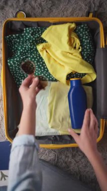 Kadın, içine giysi, su şişesi ve kulaklık doldurulmuş sarı bir bavul hazırlıyor, üstüne de kapalı bir ortamda seyahat hazırlıklarını gösteren bir hasır şapka takıyor.