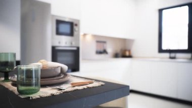 Modern mutfak sahnesinde tabaklar, çatal bıçaklar ve yeşil gözlüklerle süslenmiş siyah bir tezgah sergileniyor. Parlak, minimalist bir zemin üzerine kurulmuş ve doğal ışığın atmosferi artırmasına olanak sağlayan bir pencere var.