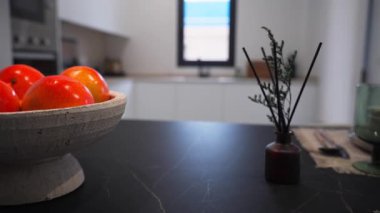 Parlak portakallarla dolu bir kase modern bir mutfağın koyu bir tezgahında duruyor. Yanında da dekoratif bir vazo ve cam süs var.