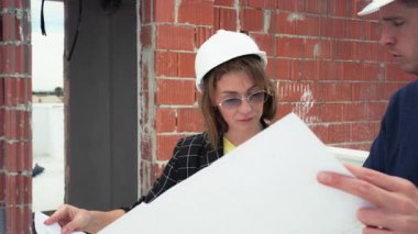 Sert başlıklı kadın inşaat işçisi, inşaat alanındaki erkek meslektaşıyla birlikte planları gözden geçirir, arka planda tuğla duvarlar ve aletler olan proje özellikleri hakkında tartışır.