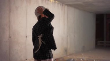 Kısmen inşa edilmiş bir binada, arka planda enkaz ve bir meslektaşı dururken el hareketleriyle el kol hareketi yapan, kareli ceketli ve kasketli bir kadın inşaat işçisi var.