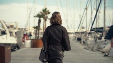 Modaya uygun siyah bir kadın marina geçidi boyunca yürüyor, bir gülümsemeyle geriye bakıyor, yatlar ve palmiyelerle çevrili, doğal güzelliklerle dolu sakin bir sahil manzarası yaratıyor.