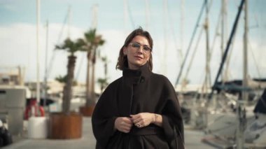 Modaya uygun siyah ceketli kısa kahverengi saçlı kadın marinada poz veriyor, arka planda yatlar ve palmiye ağaçları, parlak bir gökyüzü altında canlı bir kıyı sahnesi yakalıyor.