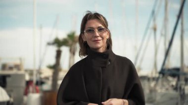 Açık kahverengi saçlı, gözlüklü ve koyu renk ceketli bir kadın, tekneler ve palmiye ağaçları parlak bir gökyüzünün altında manzaralı bir zemin oluştururken, gözlüğünü bir marinanın yanında rahatça ayarlıyor.