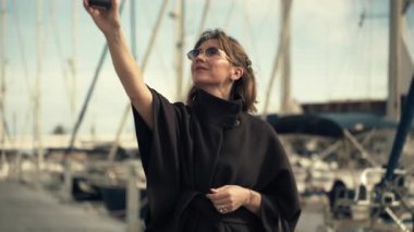 Siyah pançolu kadın marinada poz veriyor, akıllı telefonuyla selfie çekiyor, palmiye ağaçları ve yatlarla çevrili, canlı bir sahil atmosferi yaratıyor.