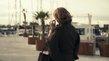 Kısa kahverengi saçlı bir kadın marinada geziniyor, elinde akıllı bir telefon, arka planda palmiye ağaçları ve botlar var, sakin sahil çevresini geliştiriyor.