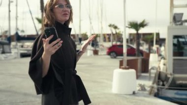 Havalı siyah elbiseli bir kadın marina boyunca yürüyor, elinde akıllı bir telefon, arka planda botlar ve palmiye ağaçları ile, hareketli bir kıyı sahnesi yaratıyor.