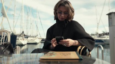 Siyah pelerinli açık kahverengi saçlı bir kadın marinanın yanındaki bir masada oturuyor, akıllı telefonuyla meşgul, teknelerle çevrili ve açık gökyüzü, açık hava ortamını güzelleştiriyor.