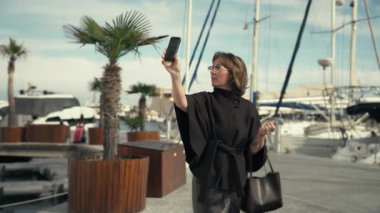Siyah elbiseli orta boylu bir kadın marinada akıllı telefonuyla poz veriyor, yat ve palmiye ağaçlarına selfie çekiyor, canlı bir deniz manzarası yansıtıyor.