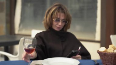 Orta boylu, kahverengi şallı bir kadın, açık hava masasında akıllı telefonuna odaklanırken bir bardak şaraptan zevk alıyor. Mavi masa örtüsünün üzerinde bir tabak ve bir sepet ekmek var.