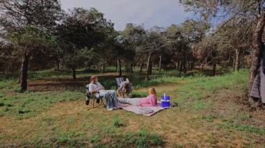 Aile üyeleri bir orman açıklığında bir battaniyenin üzerinde toplanmış ağaçlar ve yeşilliklerle çevrili bir şekilde sohbet ve rahatlama içinde birlikte huzurlu bir açık hava pikniğinin tadını çıkarıyorlar.
