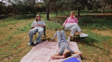 Üç genç kadın çizgili bir battaniyenin üzerinde sakin bir anın tadını çıkarırken, diğerleri ağaçların ve çimenli arazinin önünde, sandalyelerde oturuyorlar.