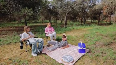 Üç genç kadın ormanda gündelik pikniğin keyfini çıkarırken, biri battaniyenin üzerinde yatarken diğerleri yemyeşil ve ağaçlarla çevrili katlanan sandalyelerde huzurlu bir atmosfer yaratıyorlar.