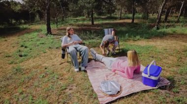Üç genç birey, çimenli bir alanda piknik battaniyesinde bir kişi yatarken, diğerleri katlanan sandalyelerde oturup sıcak güneş ışığı ve doğal çevrenin tadını çıkarıyorlar.