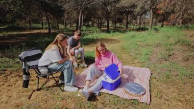 Genç arkadaşlar ormanda piknik yapmak için toplanıyorlar. Bir kadın soğutucuyu açarken, diğerleri ağaçların altındaki çizgili bir battaniyeye oturmuş doğal bir şekilde izliyor.