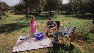 İki kadın ve bir oğlan güneşli bir ormanda çizgili bir battaniyeye oturmuş, serinletici ve içeceklerle pikniğe gidiyorlar, yemyeşil ve sakin bir açık hava ortamında ağaçlarla çevrili.