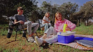 Üç genç arkadaş güneşli bir açık hava ortamında piknik yapmak için toplanırken, pembe kazaklı bir kadın soğutucuyu açarken, diğer ikisi de çimenli alanda katlanan sandalyelerde dinleniyor.