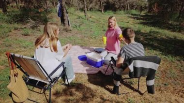 Üç genç arkadaş güneşli bir ormanda piknik yapmak için toplanırken, bir kadın battaniyede oturmuş, etrafı ağaçlar ve yemyeşil yeşilliklerle çevrili bir şekilde soğutucudan içecek servisi yapıyor.