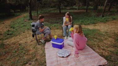 Üç genç arkadaş ormanda bir piknik battaniyesinde oturmuş, ağaçlar ve otlarla çevrili sarı bardaklardan içecekleri yudumlarken, dinlenmek için sakin bir açık hava ortamı yaratıyorlar.