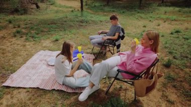 Pembe kazaklı bir kadın ve çizgili bir battaniyenin yanındaki katlanabilir sandalyeye oturmuş renkli bardaklar tutan iki çocukla, çimenli bir ortamda piknik yaparken üç kişi rahatlıyor.