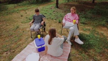 Çimenli bir alanda piknik yapmak için toplanan bir grup çocuk, daha serin ve renkli bardaklarla bir battaniyenin üzerinde oturuyor, ağaçların gölgesi altında birbirleriyle etkileşime giriyor ve eğleniyorlar.