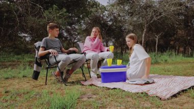 Üç çocuk dışarıda yeşil bir alanda piknik yapmak için toplanıyor, ikisi sandalyede, biri battaniyede oturuyor, soğutucuda içki ve atıştırmalıkları paylaşıyor, ağaçlar ve çimlerle çevrili.