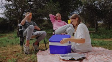 Üç genç arkadaş, katlanır sandalye ve battaniyenin üzerine oturmuş, güneşli havanın ve doğal çevrenin tadını çıkarırken içki ve atıştırmalıkları paylaşıyor.