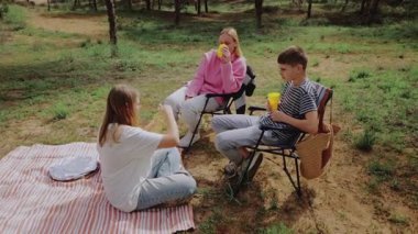 Üç genç arkadaş ormanda bir piknik battaniyesinde oturmuş, yemyeşil ağaçlar ve yemyeşil ağaçlarla çevrili iken içeceklerini ve atıştırmalıklarını paylaşıyor, neşeli ve rahat bir açık hava ortamı yaratıyorlar.