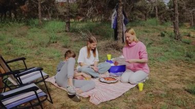 Çimenli bir alanda piknik yapmak için toplanan aile, iki çocuğa yemek servisi yapan bir kadınla birlikte bir battaniyenin üzerinde, ağaçlarla çevrili ve açık havada rahat bir ortam yaratmak için.
