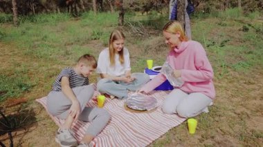 Bir kadın ve iki çocuğun yer aldığı aile pikniği sahnesi bir battaniyenin etrafında toplandı, çimenli bir alanda pizza ve içecekleri paylaştı, ağaçlar doğal bir zemin oluşturdu ve açık havayı renklendiren renkli bardaklar sundu.