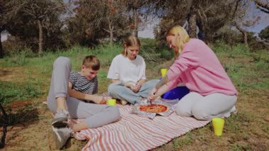 Bir grup çocuk ve kadın piknik yapmak için dışarıda toplanıyor, güneşli bir parkta, ağaçlarla çevrili bir battaniyede pizza paylaşıyorlar, renkli bardaklar ve neşeli atmosferi yükselten günlük giysilerle.