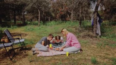 Bir grup çocuk ve bir kadın, ağaçlarla çevrili çimenli bir alanda, bir battaniyede pikniğe katlanmış bir sandalye ve yakınlarda bir soğutucuyla birlikte, rahat bir açık hava ortamı yaratıyor.