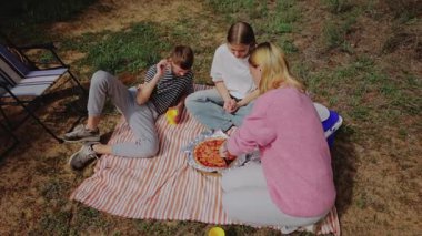 Çocuklar ve kadınlar çimenli bir alanda çizgili bir battaniyenin etrafında toplanarak gümüş tepside pizza paylaşıyorlar, arka planda katlanan sandalyeler ve soğutucu ile rahat bir açık hava yaratıyorlar.