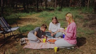 Üç genç arkadaş ormanda piknik yapmak için toplanıyor, battaniyenin üzerinde yemek ve kahkaha paylaşıyorlar, yakınlarda katlanır sandalyeler ve ağaçlar sakin bir açık hava yaratıyor.