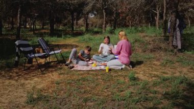 Bir grup çocuk ve kadın, ağaçların altındaki çimenli bir alanda piknik battaniyesine oturmuş, yiyecek ve içecekleri, katlanır sandalyeleri ve güneşli açık havada serin bir hediyeyi paylaşıyorlardı.