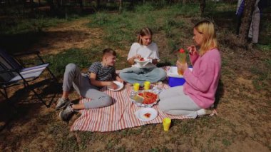 Aile, ormanda çizgili bir battaniyede piknik yapmak için toplandı. Çocuklar çimlerin üzerinde oturmuş yiyecek ve içeceklerin tadını çıkarırken, anne de yanlarında duran soğutucudan yiyecek servisi yapıyordu.