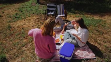 Üç kadın, çimenli bir alanda çizgili bir battaniyede toplanarak yemek ve içeceklerle pikniğin keyfini çıkarırken, iki sandalye de parlak güneş ışığının altında yakınlarda yer alıyor.