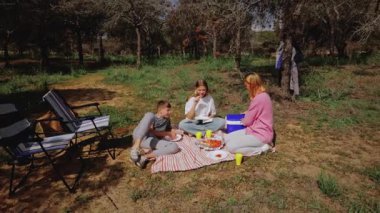 Çocuklar battaniye ve içecekleri paylaşırken, yetişkinler ağaçların altındaki katlanan sandalyelerde rahatlarken aile güneşli bir açık alanda piknik yapmak için bir araya geldi.