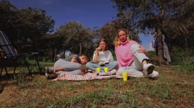 Genç arkadaşlar yeşil bir parkta piknik yapmak için toplanıyorlar, bir battaniyenin üzerinde oturuyorlar ve güneşli havanın, ağaçların ve otların doğal ortamının tadını çıkarırken yiyecek ve içeceklerini paylaşıyorlar.