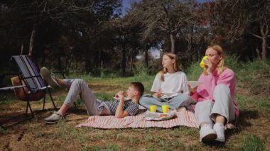 Aralarında bir kadın ve iki çocuğun da bulunduğu üç kişi, ağaçlar ve yeşilliklerle çevrili renkli fincanlar ve yiyeceklerle bir pikniğin tadını çıkarırken, ormanlık bir alanda çizgili bir battaniyenin üzerinde oturuyorlar.