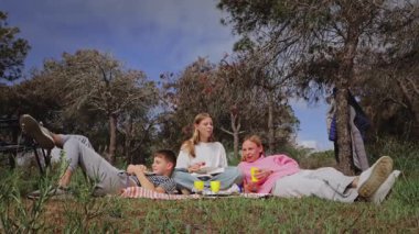 Üç genç arkadaş ormanlık bir alanda piknik battaniyesinde uzanıyorlar. Açık havada huzurlu bir çevrede ağaçlar ve yeşilliklerle çevrili olarak birbirlerinin yanında yiyecek ve içeceklerini paylaşıyorlar.